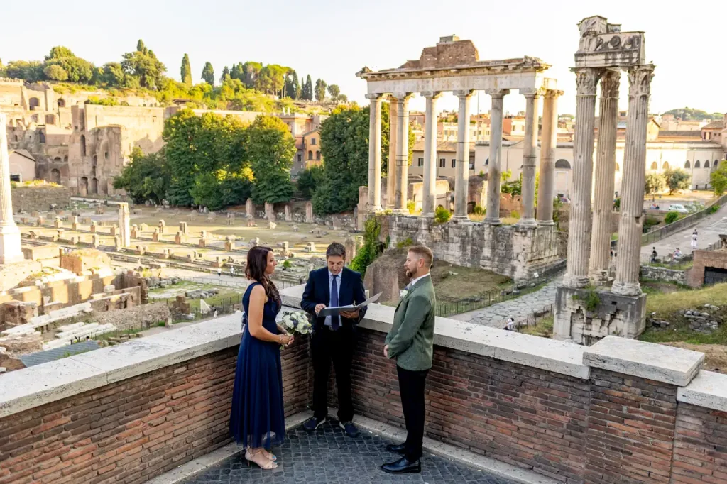 A couple standing with a celebrant during an outdoor wedding ceremony against the backdrop of the ancient ruins and columns of the historic imperial fora.