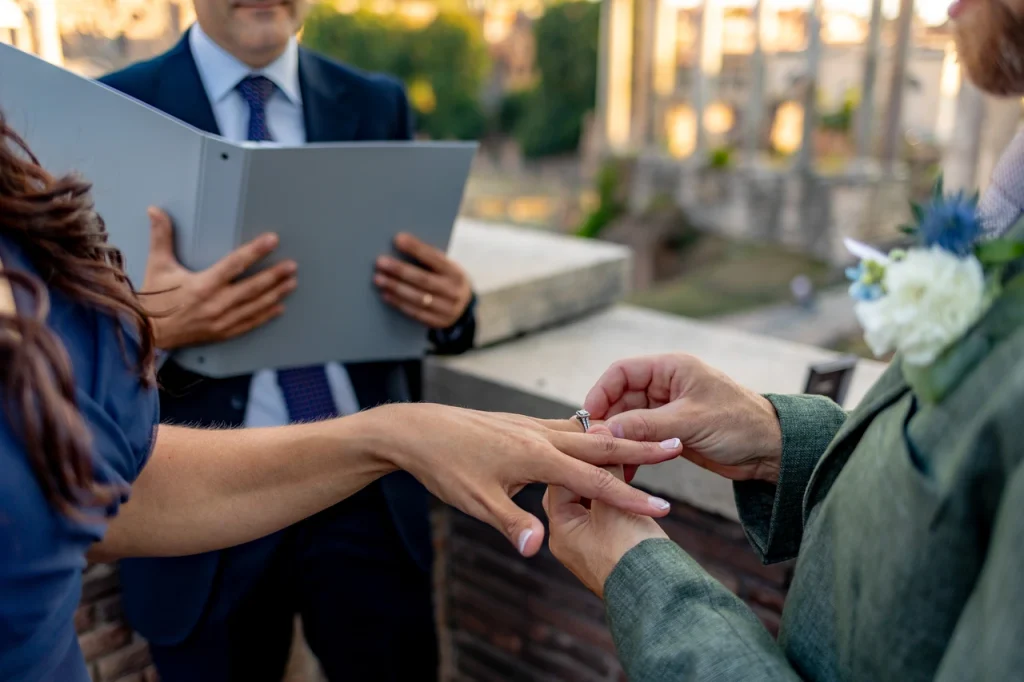 One person places a ring on another's finger during an outdoor ceremony, with a celebrant holding a book in the background.