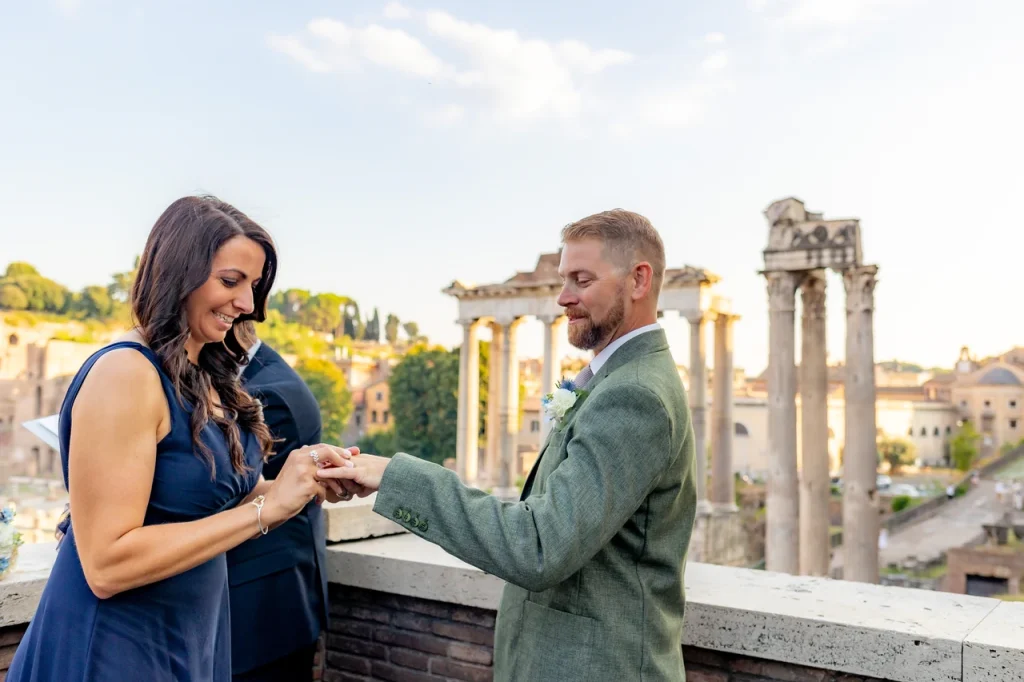 A couple exchange rings on a balcony overlooking ancient roman ruins, with clear skies and historic columns in the background.