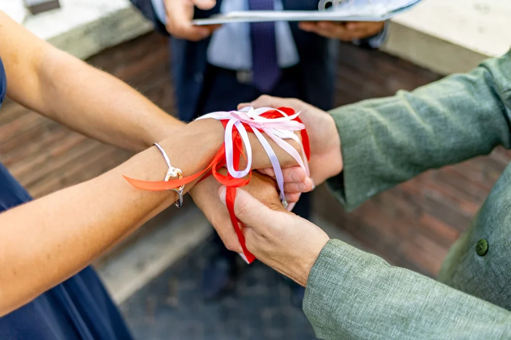 Close-up of hands during an elopement ceremony, with red and white ribbons tied around the couple's wrists and a celebrant holding a document in the background.
