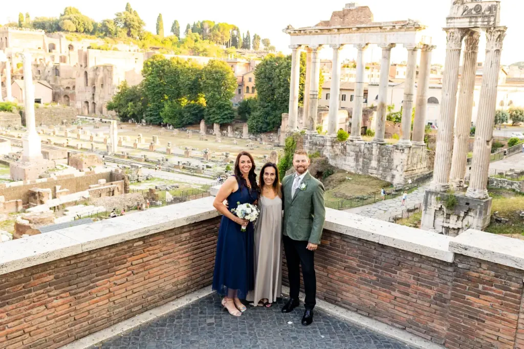 Elopement italy. The bride and groom pose on a terrace with ancient ruins and columns in the background, together with wedding planner simona celani.