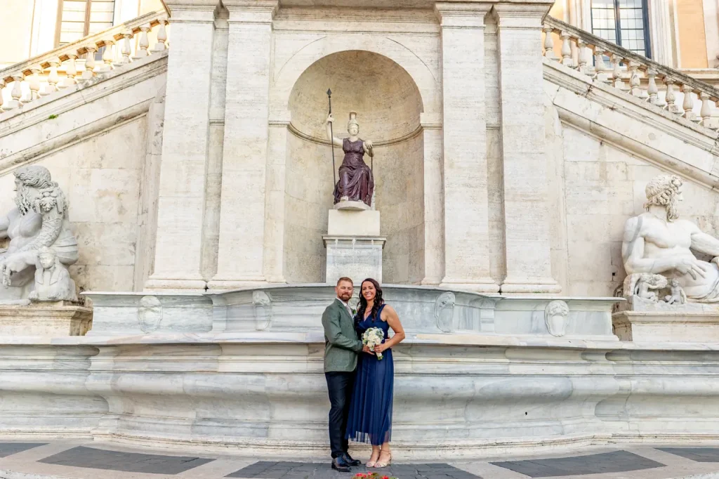 A couple poses in front of a historic stone staircase and statues, with the woman holding a bouquet of flowers, while a statue of the “goddess rome” stands majestically in the background.