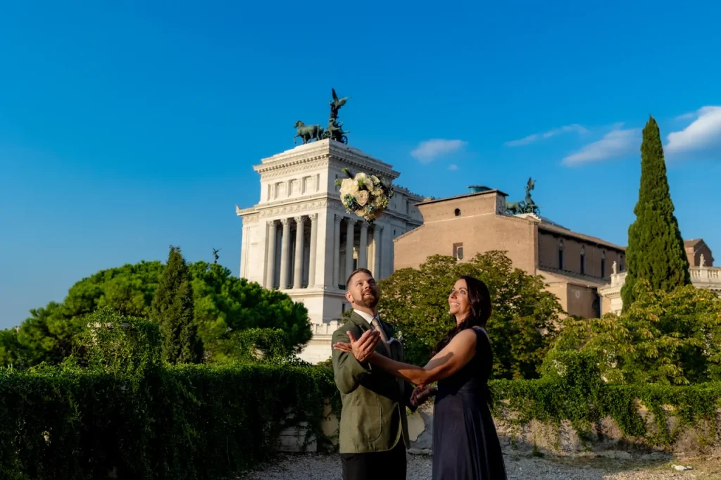A man and a woman stand together smiling, in the background the historic altar of the fatherland with columns and statues, under a clear blue sky.