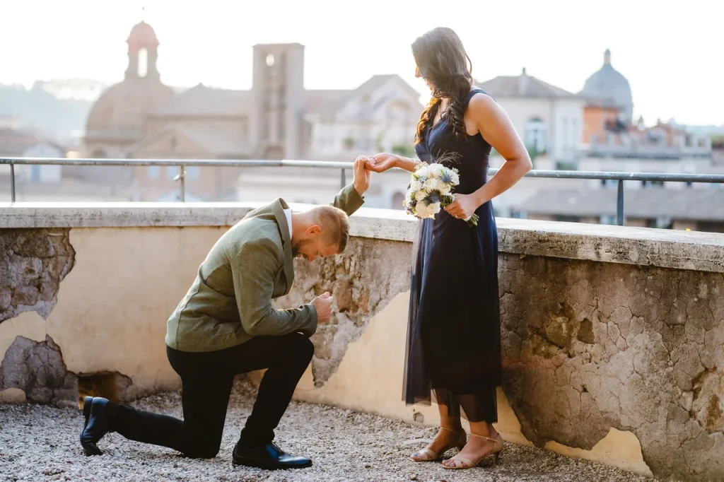 A man is kneeling and asking for the hand of a woman holding a bouquet, on a rooftop with city buildings in the background.