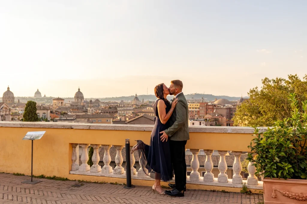 A couple kisses on a terrace, savoring the view of rome with its domed buildings and clear sky.