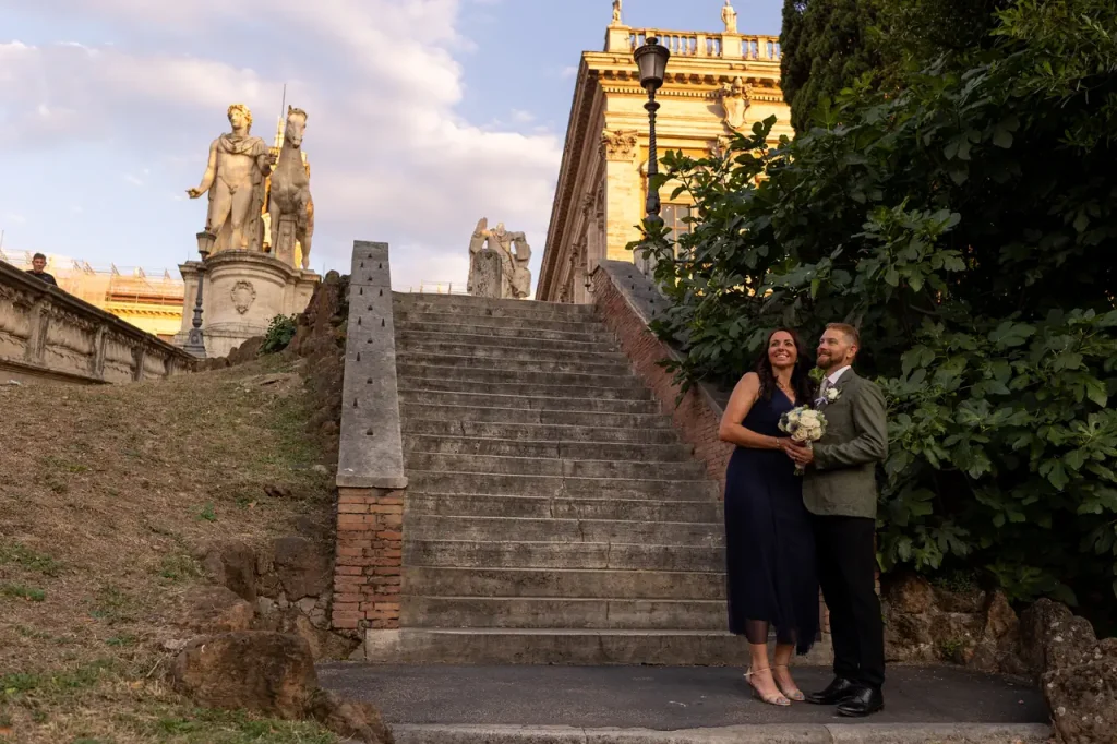 A couple is standing at the foot of stone steps next to a historic building, holding a bouquet of flowers, while statues can be seen in the background.