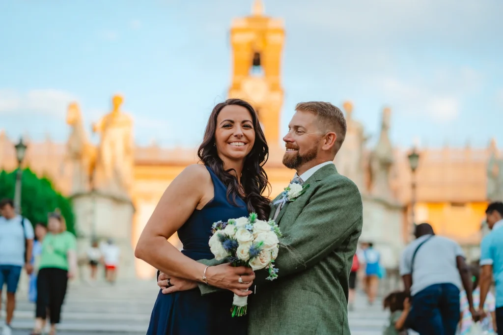 A couple poses together, with the woman holding a bouquet. They are standing in front of a historic building with statues and people in the background.