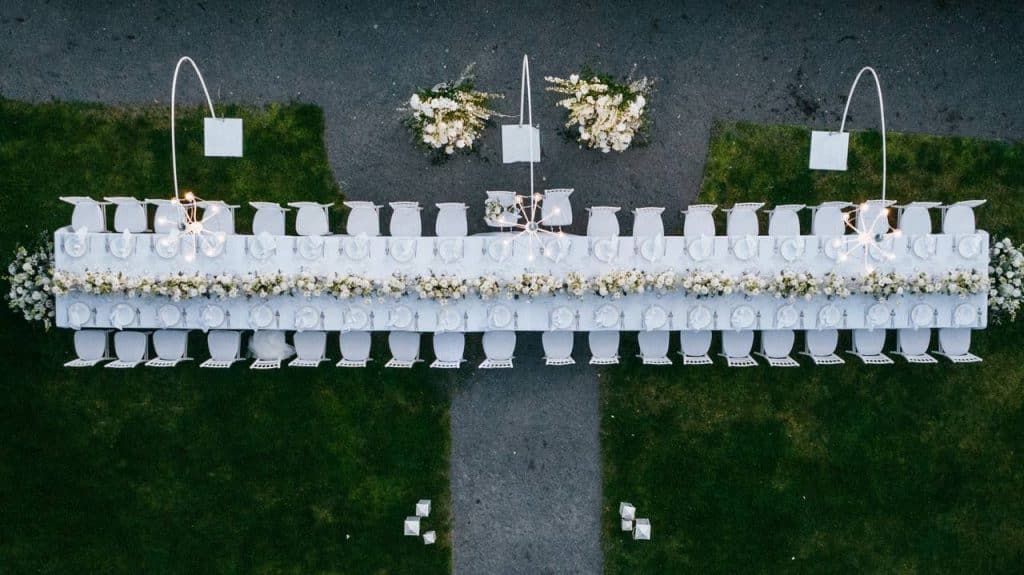 Aerial view of a long outdoor table set up with white chairs and floral decorations, arranged symmetrically on a lawn.