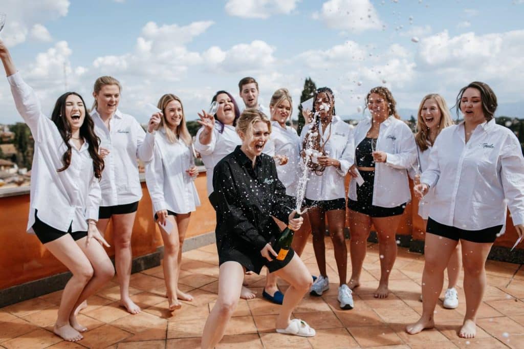 A group of people, mostly wearing oversized white shirts, are standing on a terrace. A person in the centre uncorks a bottle of champagne, with liquid pouring out of it. The sky is partly cloudy.