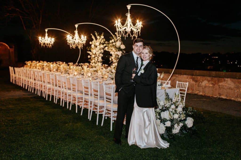 In the evening, a married couple smiles in front of a long outdoor dining table, elegantly set with white chandeliers and chairs, surrounded by flower arrangements.