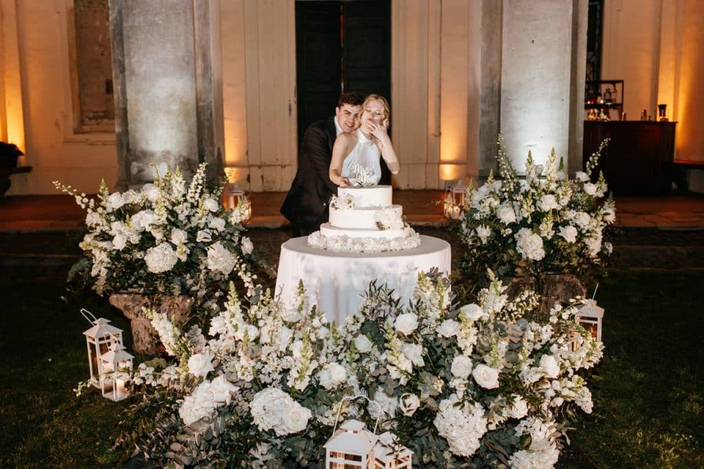 A couple cuts the wedding cake surrounded by white flower arrangements and lanterns in an outdoor setting.