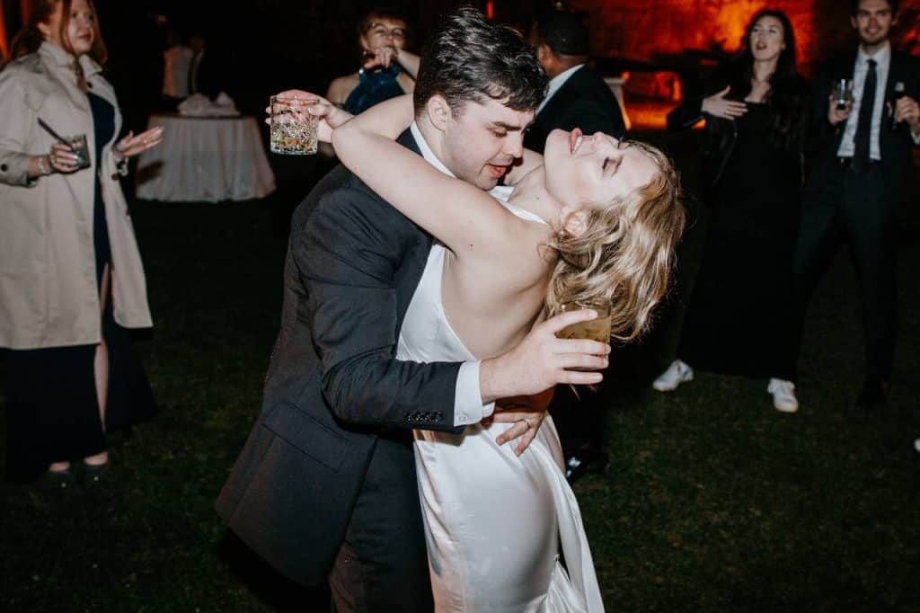 A couple dances tightly at an outdoor event, holding drinks. Several people in the background seem to be socialising and enjoying the evening.