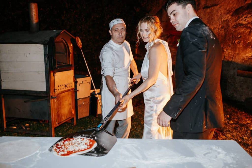A chef helps the bride to bake a pizza in an outdoor oven, under the eyes of the groom.