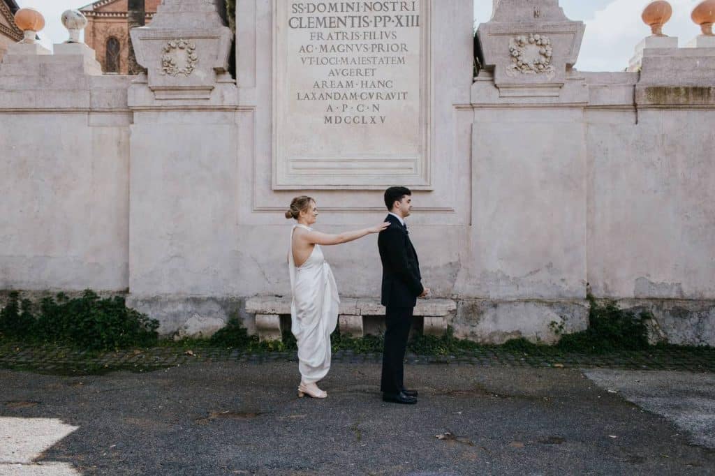 A woman dressed in white is standing behind a man dressed in black, touching his shoulder, in front of an engraved stone wall.
