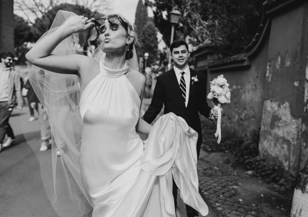The bride in a halter dress poses playfully with sunglasses while holding her veil. The groom follows her, holding the bouquet and his suit. They walk outside with spectators in the background. Black and white photo.