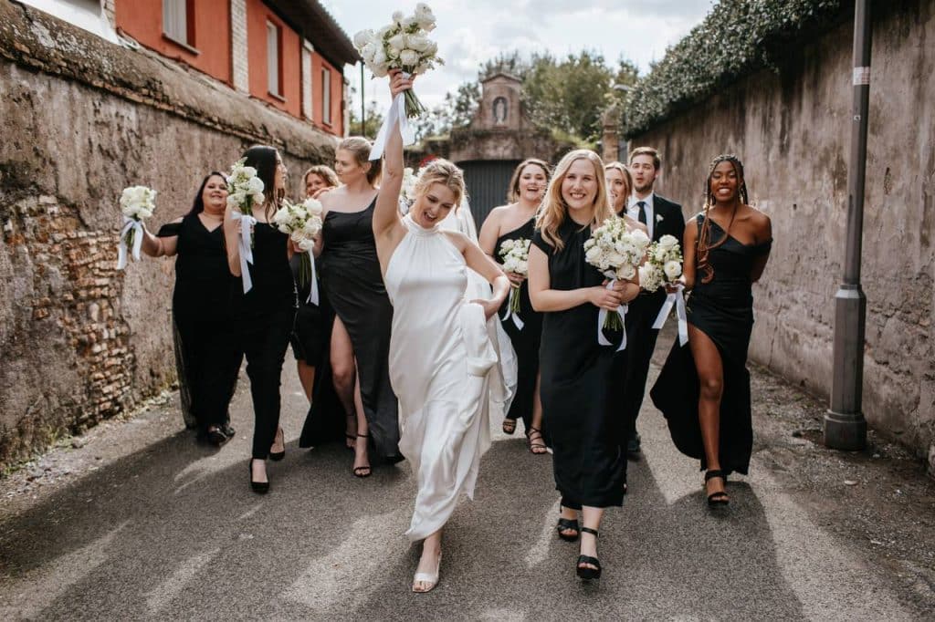 A joyful bride in a white dress and bridesmaids in black dresses walk and celebrate outdoors, holding bouquets of white flowers.