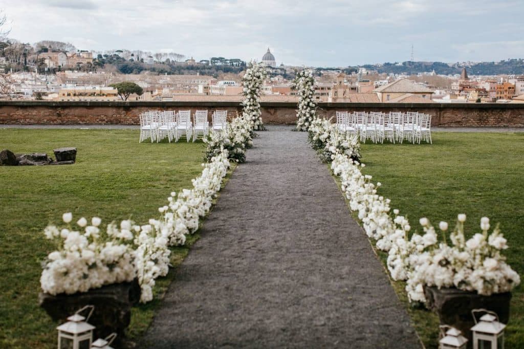 Terrazza studi romani's outdoor wedding venue features a path lined with white flowers leading to rows of elegant white chairs, all surrounded by a beautiful cityscape in the background.