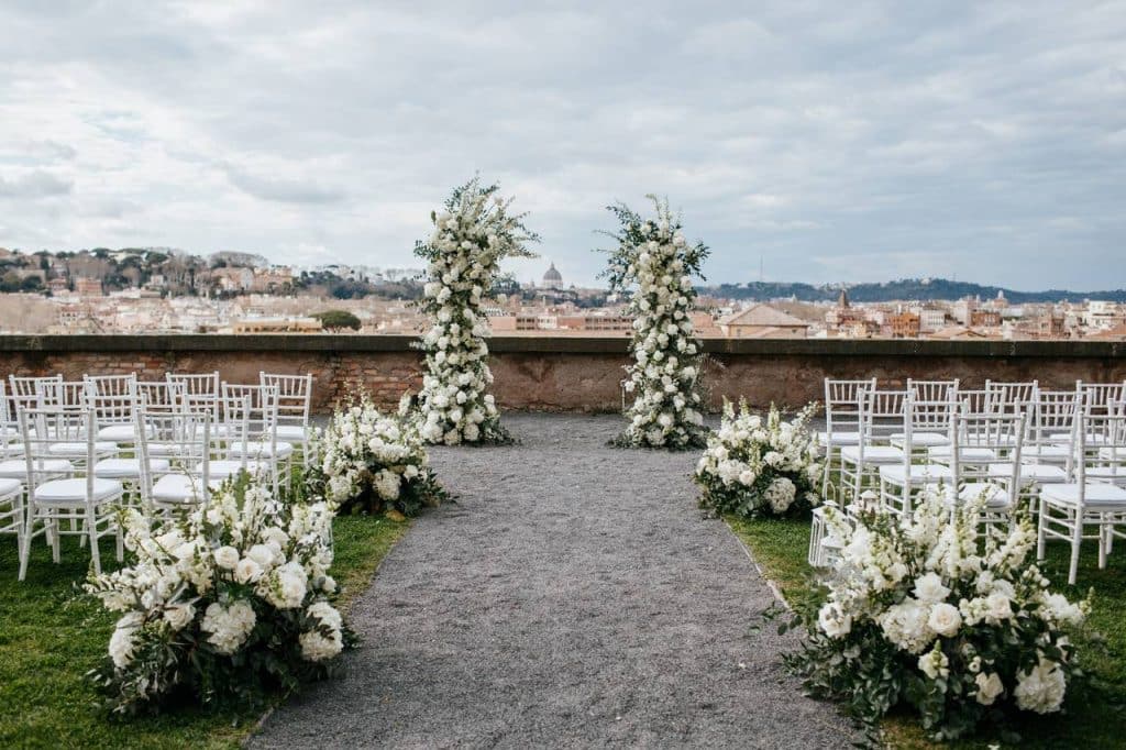 Outdoor wedding set-up with white flower arrangements and white chairs in front of two tall arches of flowers, against the backdrop of an urban landscape and a cloudy sky.