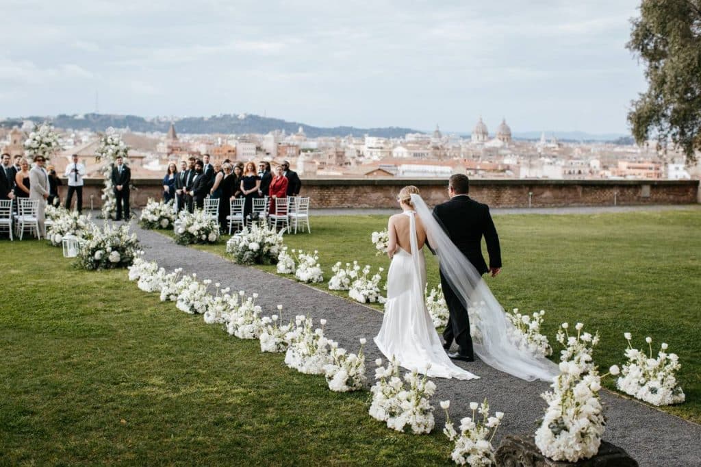 The bride and groom walk down a flower-lined outdoor aisle towards the seated guests, with a view of the city in the background.