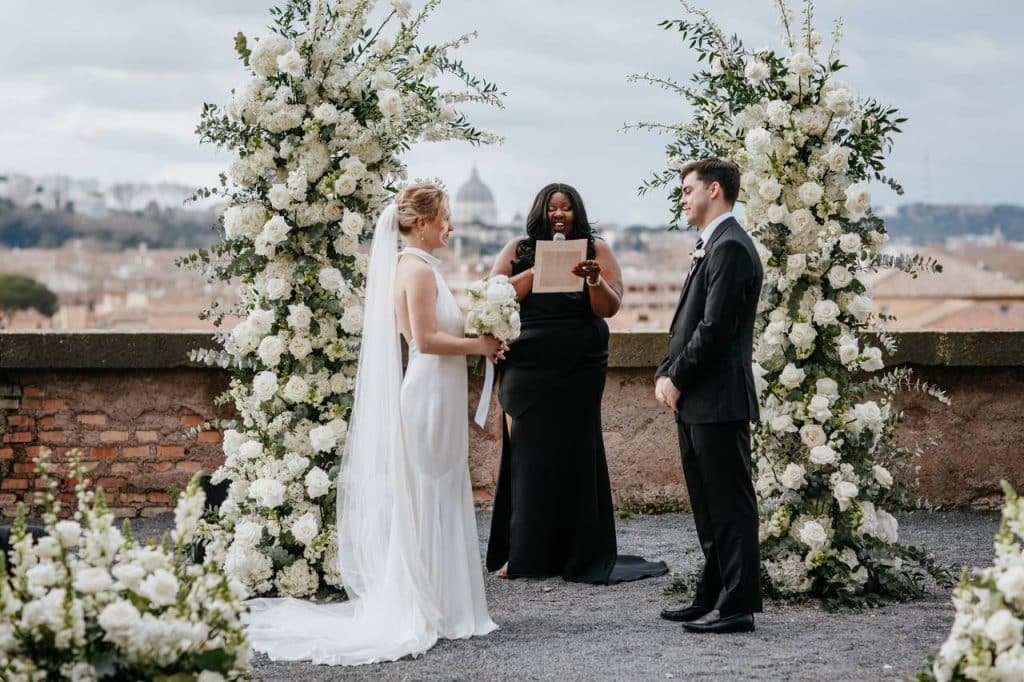 A bride and groom stand facing each other during an outdoor wedding ceremony. A celebrant stands between them, holding a paper. White floral arrangements decorate the background.