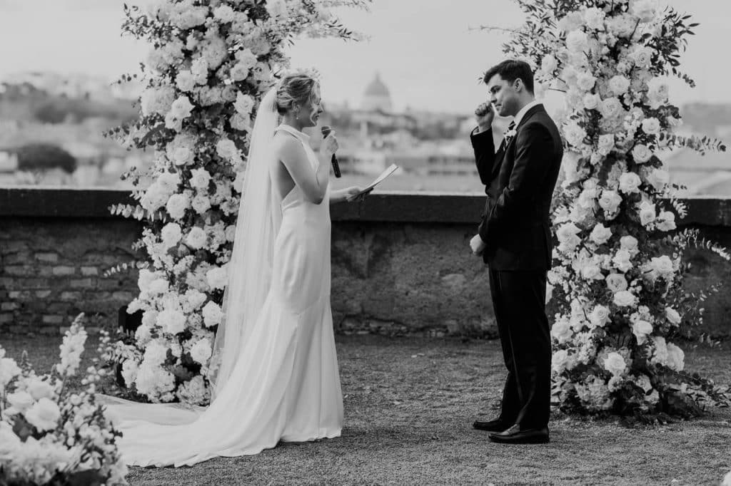 A bride and groom stand facing each other during a wedding ceremony, surrounded by floral arches, with a cityscape in the background. The bride holds notes in her hand.