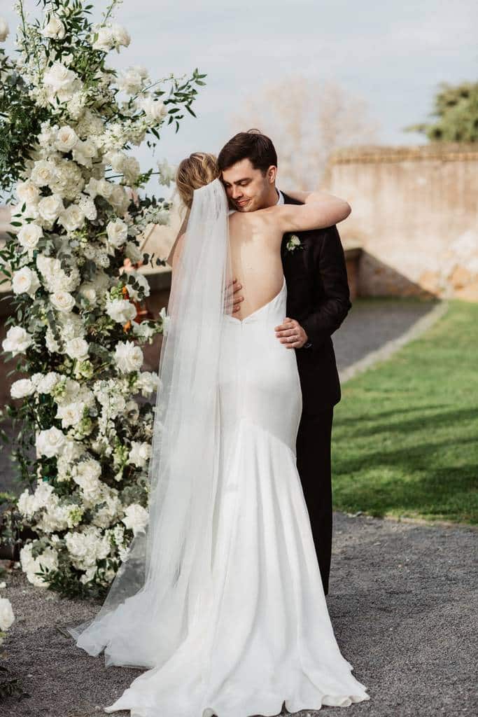 Morgan and matthew's wedding on the roman studies terrace A bride and groom embrace in front of an outdoor floral arch. The bride wears a white dress and veil, while the groom wears a black suit.