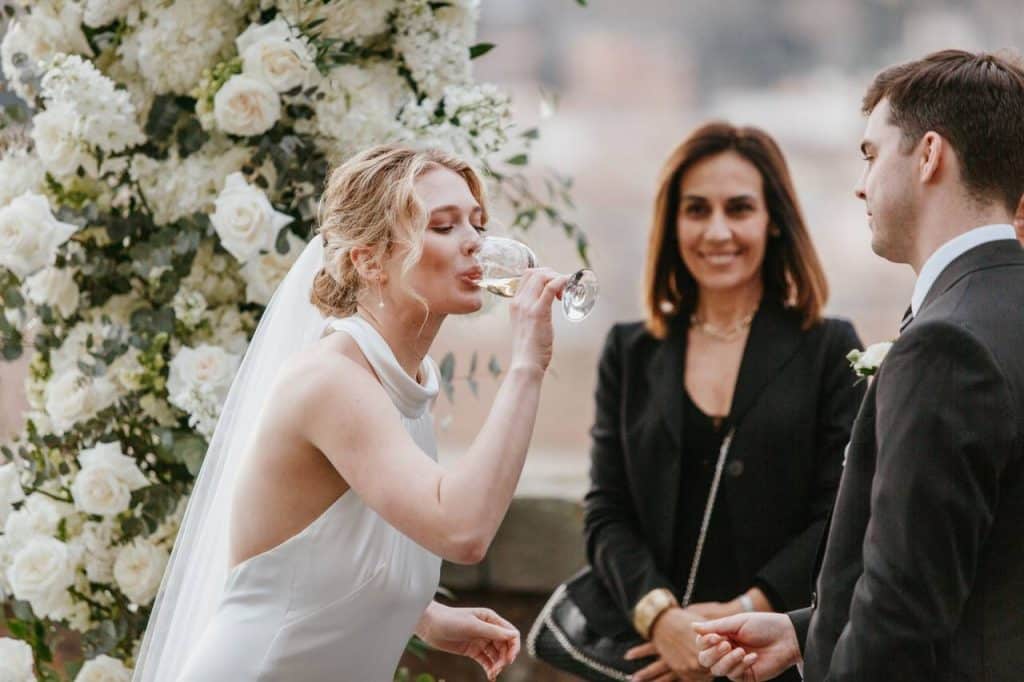 The bride sips a glass of wine during a wedding ceremony, with the groom next to her and simona celani (wedding planner in rome) standing in front of a beautifully arranged floral arch.