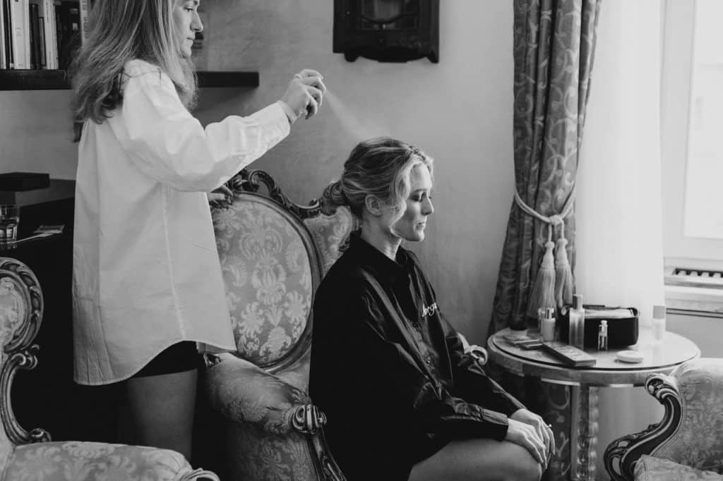 A woman styles another woman's hair in an elegantly furnished room, using a sprayer, next to a table with beauty products.