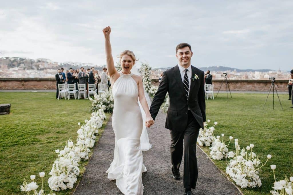 Roman terrace studios: a married couple joyfully walk down an outdoor aisle, with the bride raising her arm in celebration, surrounded by white flowers and guests in the background.