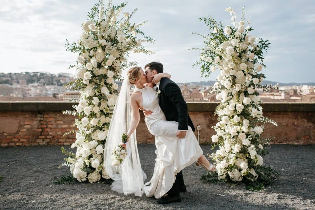 A bride and groom exchange a kiss under two floral arches, with a cityscape in the background.