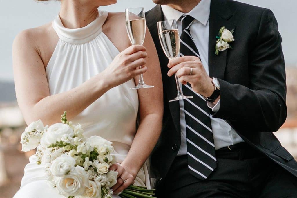 The bride in a white suit and the groom in a dark suit with a striped tie toast with glasses of champagne. The bride holds a bouquet of white flowers.