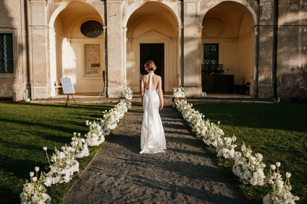 A person dressed in white walks down an outside aisle lined with white flowers, heading towards the arched entrance of a building.