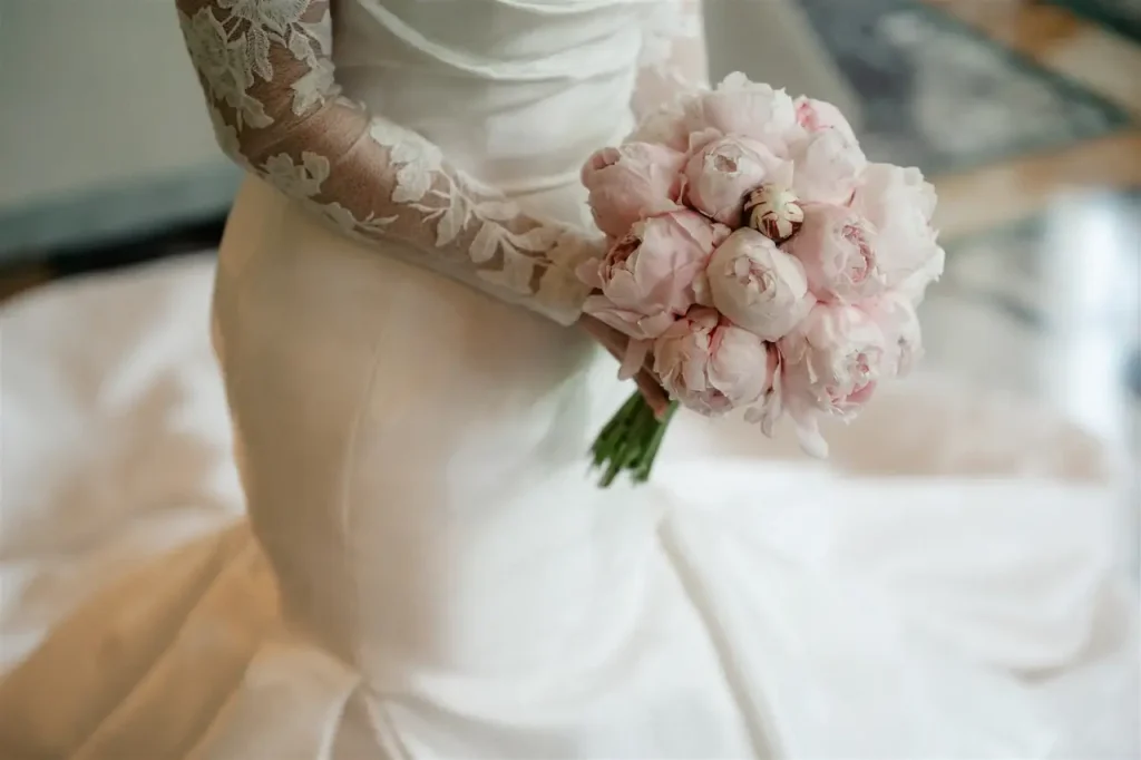 A bride in a white lace dress holds a bouquet of pale pink flowers, standing inside.
