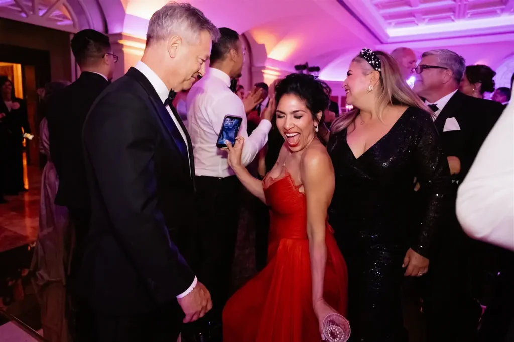 People in formal dress dancing during an indoor event, with a woman in red holding a phone and a glass, surrounded by other people in suits and elegant dresses.