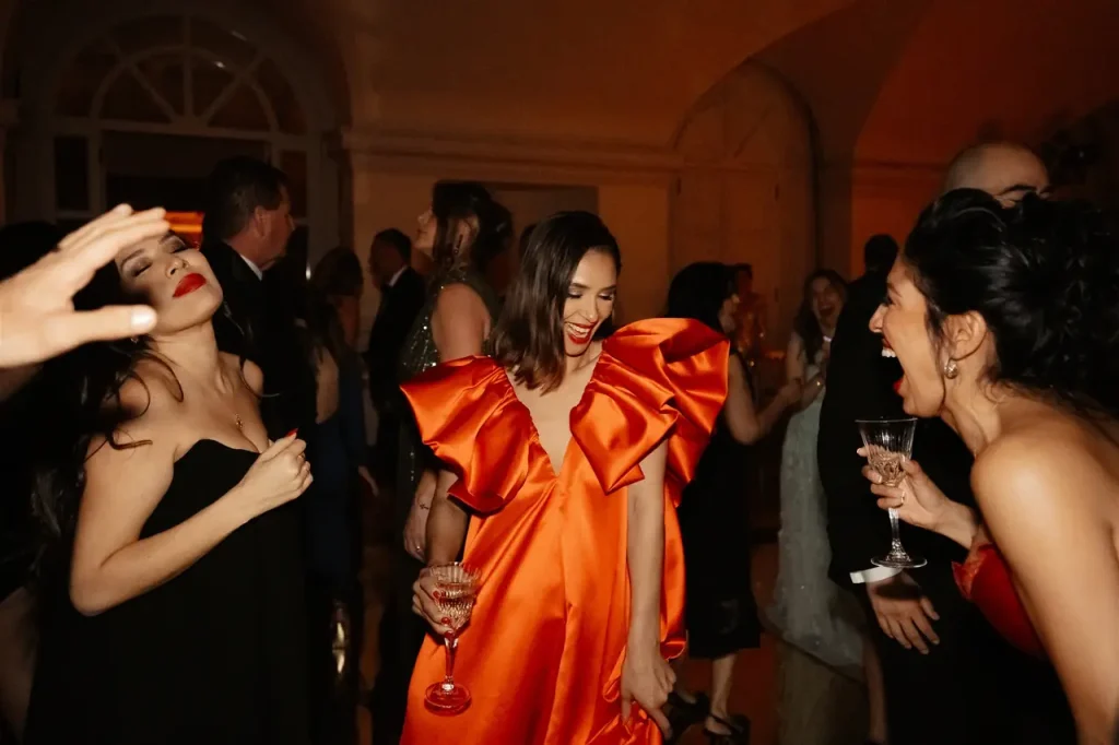 Three women, one in a bright orange dress, laugh and sip drinks at a party. Other people are in the background and the lighting is dim.