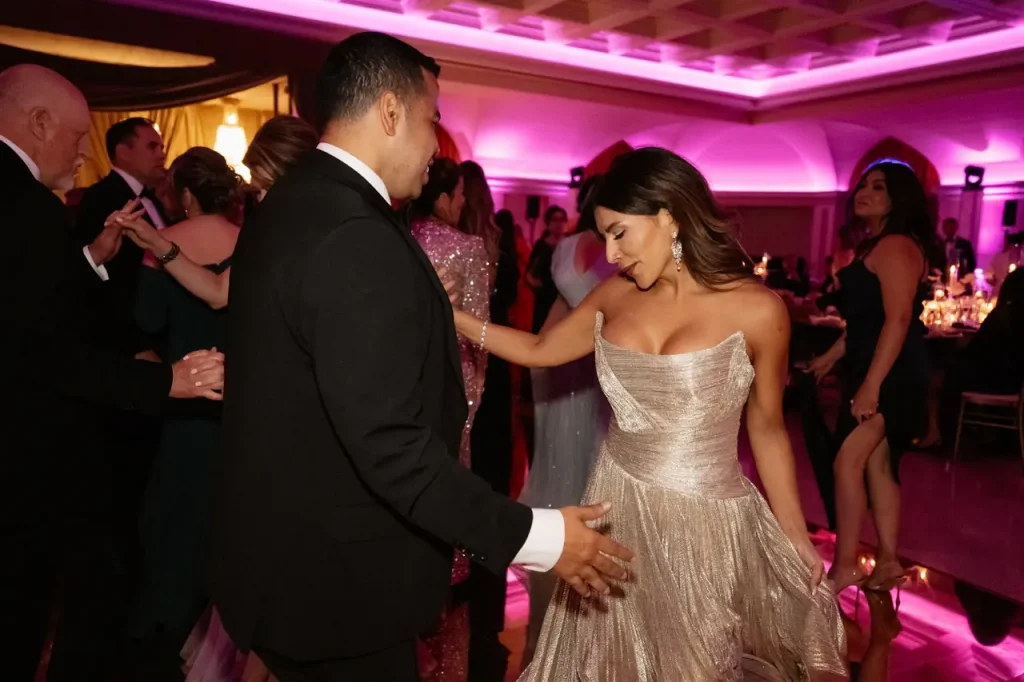 A man and a woman in formal dress dance together during an indoor event with pink lights.