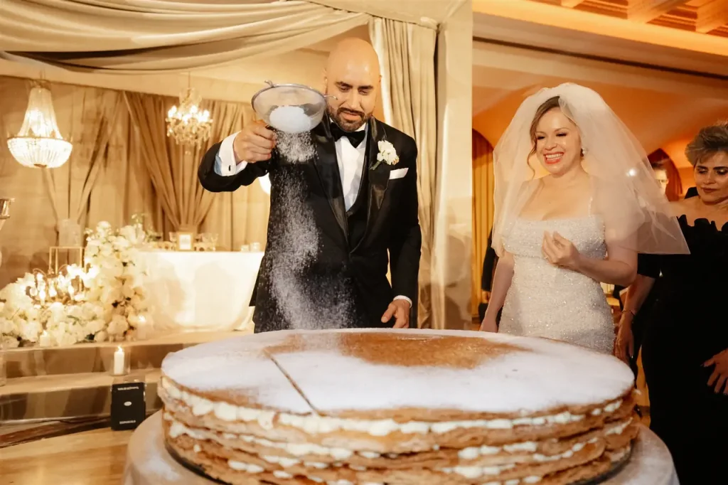 A groom sprinkles sugar on a giant layer cake while the bride and a guest look on, all in an elegantly decorated location.