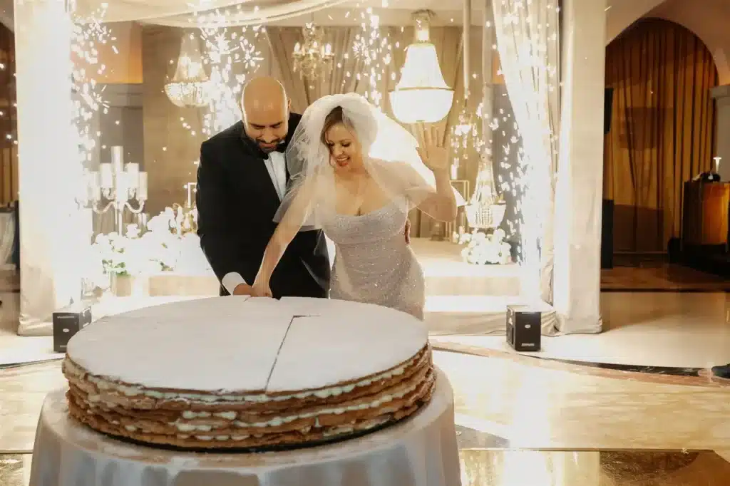 A bride and groom cut a large round cake at a wedding reception. Twinkling lights illuminate the background.