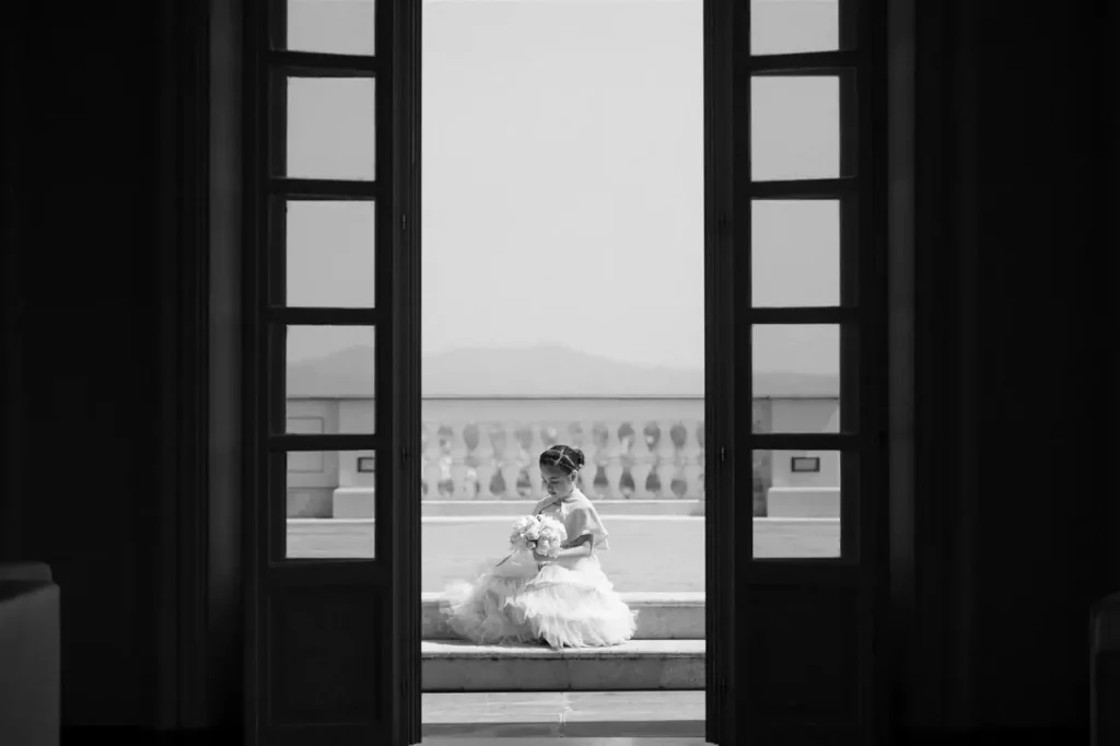 A girl dressed in white is sitting on the steps outside, framed by a large open window.