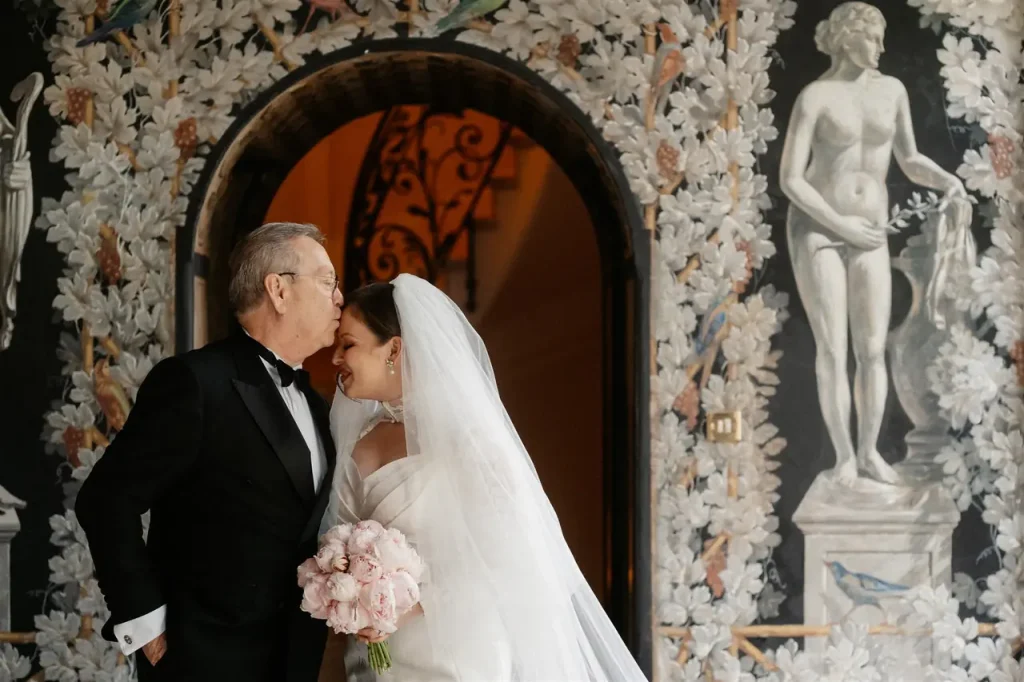 A bride holding pink roses is leaning against a groom in a dinner jacket, standing in front of a decorative arch with floral motifs and a statue.