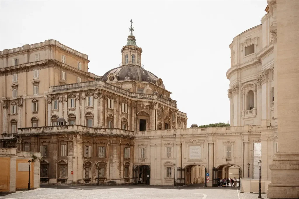 Historic baroque architecture with a domed roof and ornate façade in a sunny courtyard, characterised by arched entrances and stone details.