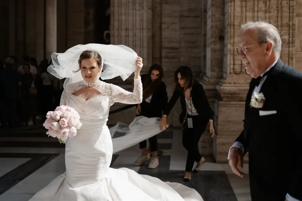 The bride with the flowers adjusts her veil while the bridesmaids adjust her dress. A man, perhaps a guest, is standing nearby. They are in a building decorated with columns.