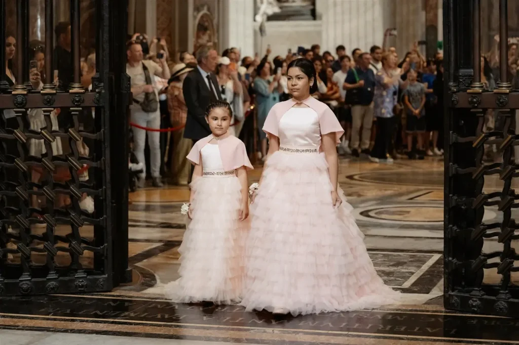 Two young girls in matching pink dresses walk down the aisle of a large ornate hall, surrounded by a crowd of onlookers.