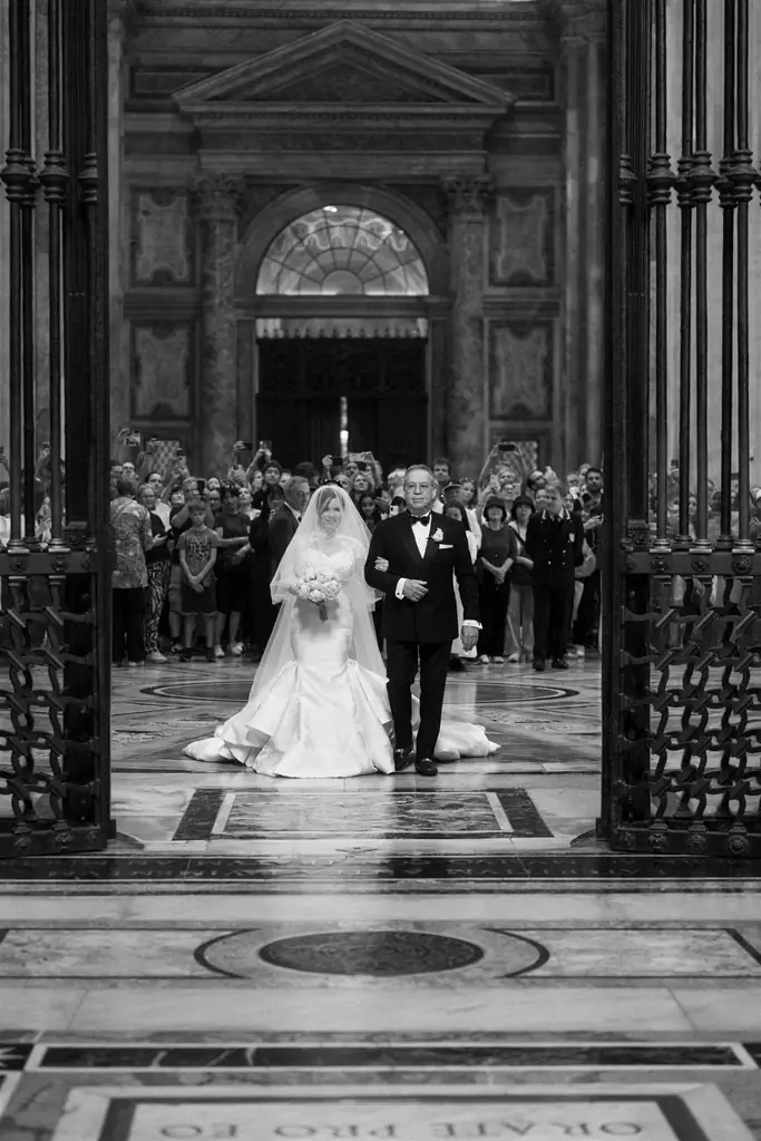Jackie and sergio's wedding, from the choir chapel to villa miani A bride in a wedding dress walks down the aisle together with an elderly man in formal dress inside a large ornate building, with spectators seated in the background.