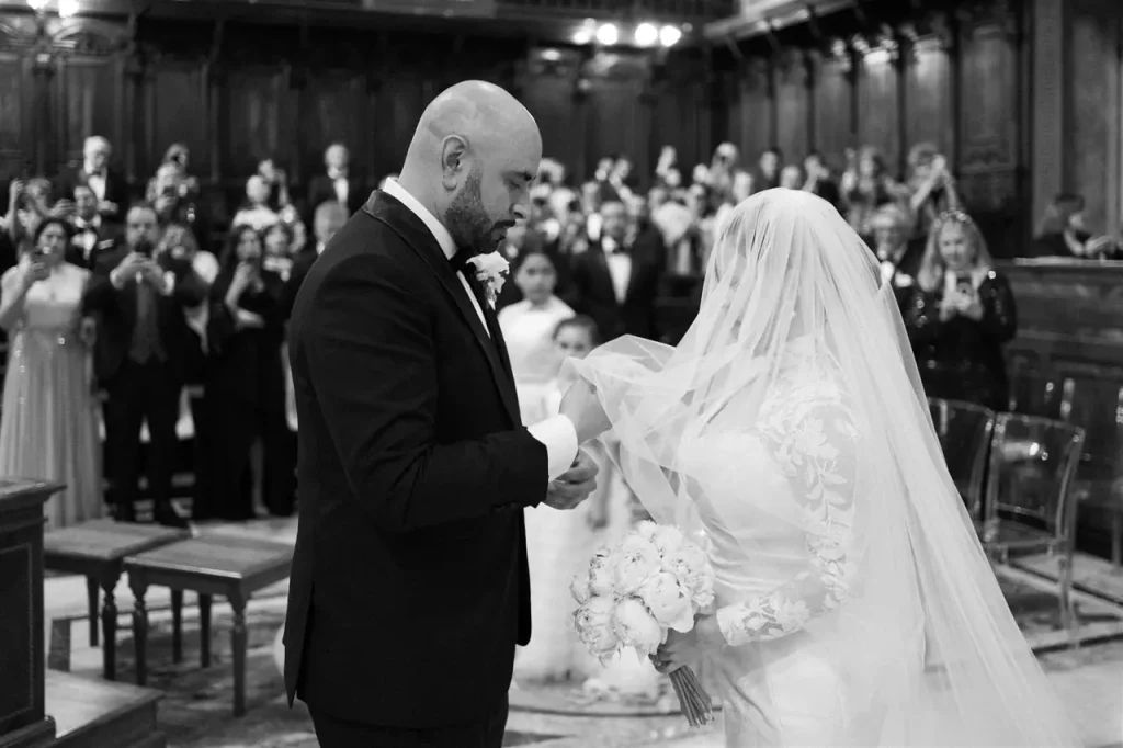 The bride and groom exchange rings in a wood-panelled room full of guests. The bride holds a bouquet and wears a veil.