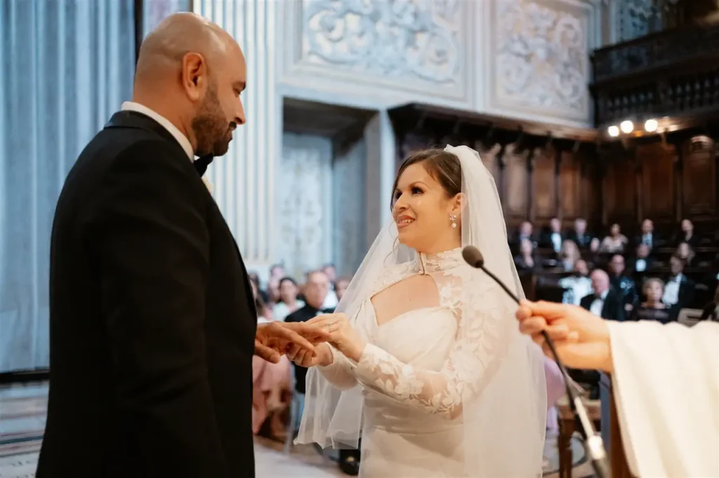 The bride and groom exchange rings during a wedding ceremony in a decorated building, with guests seated in the background.