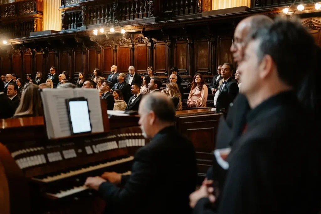 A group of people in formal dress are seated and standing inside a wood-panelled room. One person plays an organ in the foreground.