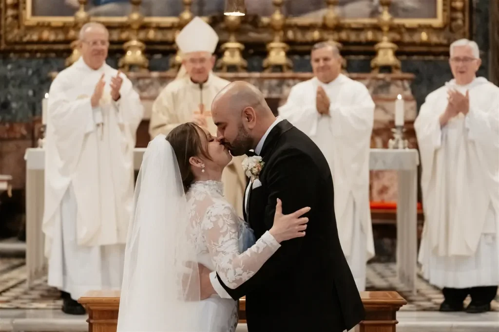 A groom and a bride kiss at the altar during the wedding ceremony. Members of the clergy in white robes stand and applaud in the background.