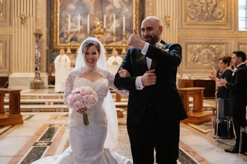 Bride and groom walk down the aisle in a church, smiling and giving a thumbs-up. The bride holds a bouquet of pink flowers, the groom in a suit. Clergy in white robes and guests applaud in the background.