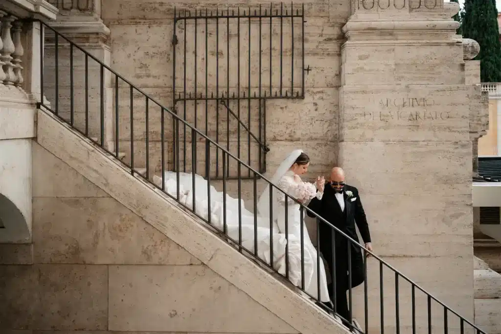 A bride in a white dress and veil descends a stone staircase with a man in a dark suit holding her hand.